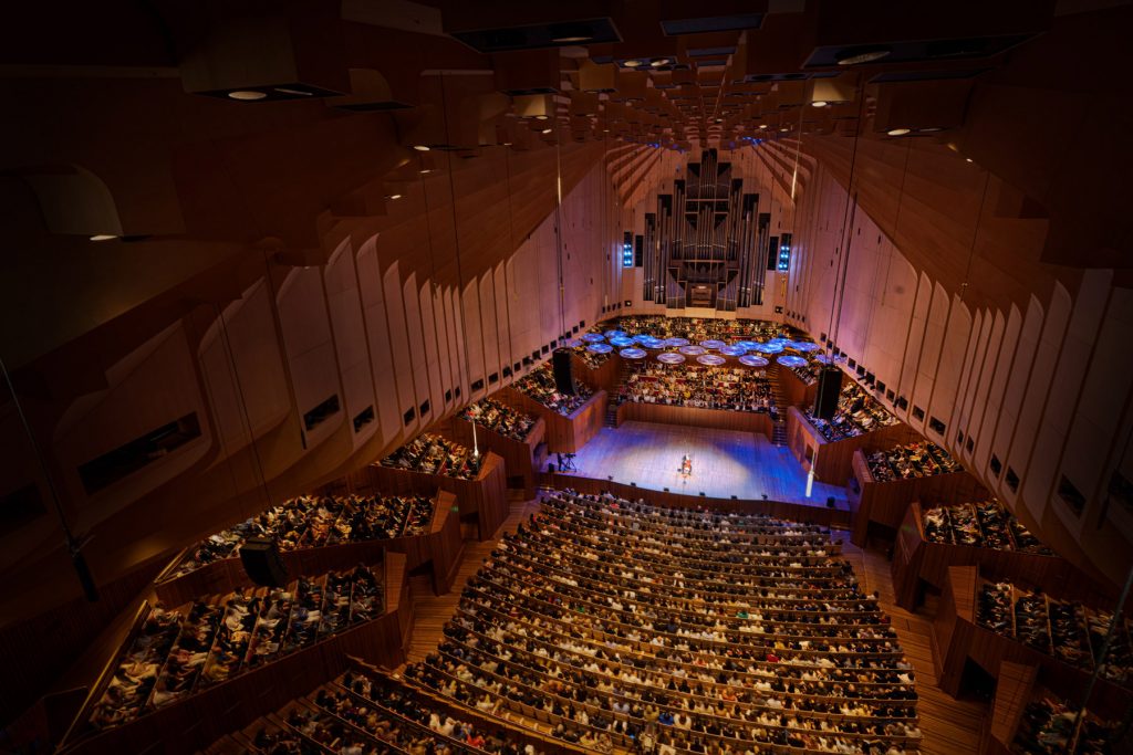 The interior of the Sydney Opera House taken from above. Cellist Yo-Yo Ma is at the center of the stage, with a white and blue spotlight on him.